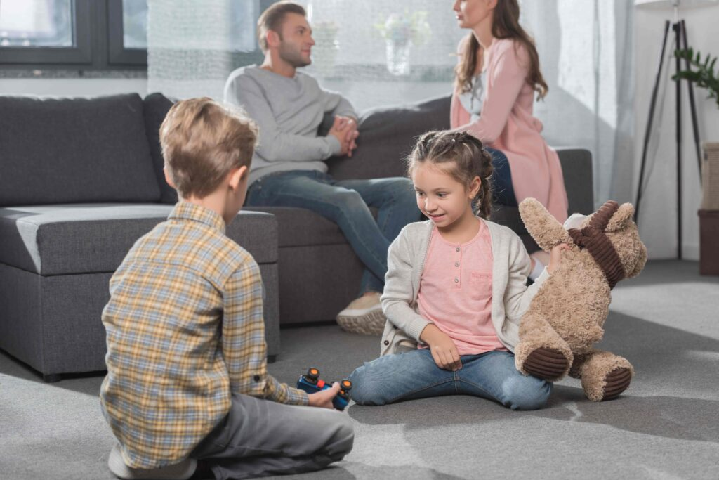 Autistic girl and neurotypical boy play together on the living room floor while parents talk in the background — reflecting family dynamics, sibling bonding, and the everyday challenges and wins of parenting a child with special needs in a supportive home environment.