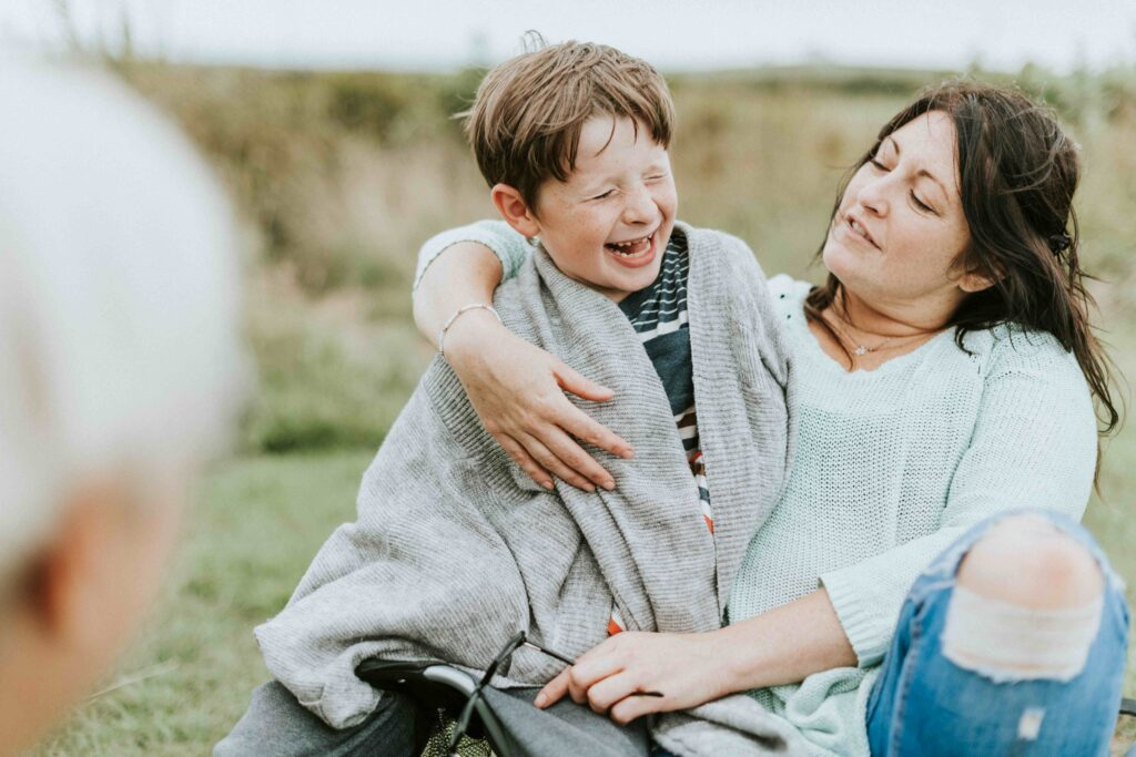 Autistic boy laughs while wrapped in a cozy blanket in his mom’s arms during a peaceful moment in nature — a heartfelt visual of the deep emotional connection, sensory regulation, and comfort provided by a dedicated mom of special needs.