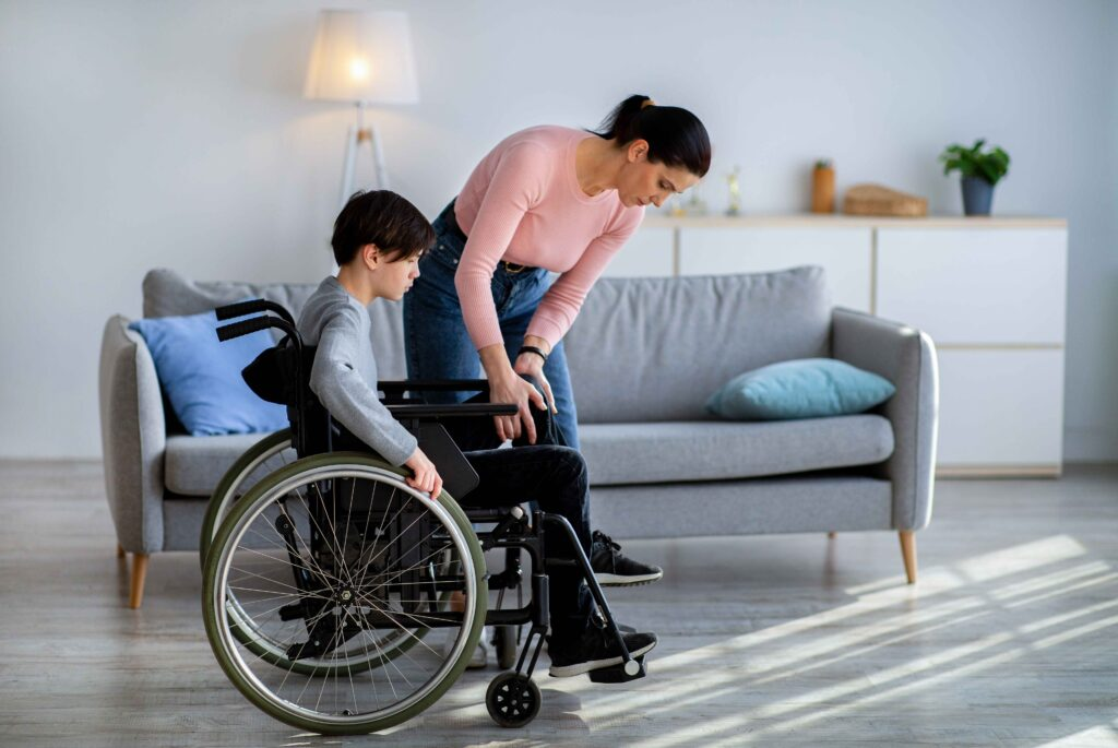 Strong and compassionate mom of special needs assists her autistic son in a wheelchair during their daily routine inside a warm home setting — a powerful visual of adaptive caregiving, mobility support, and raising a child with physical and developmental disabilities.