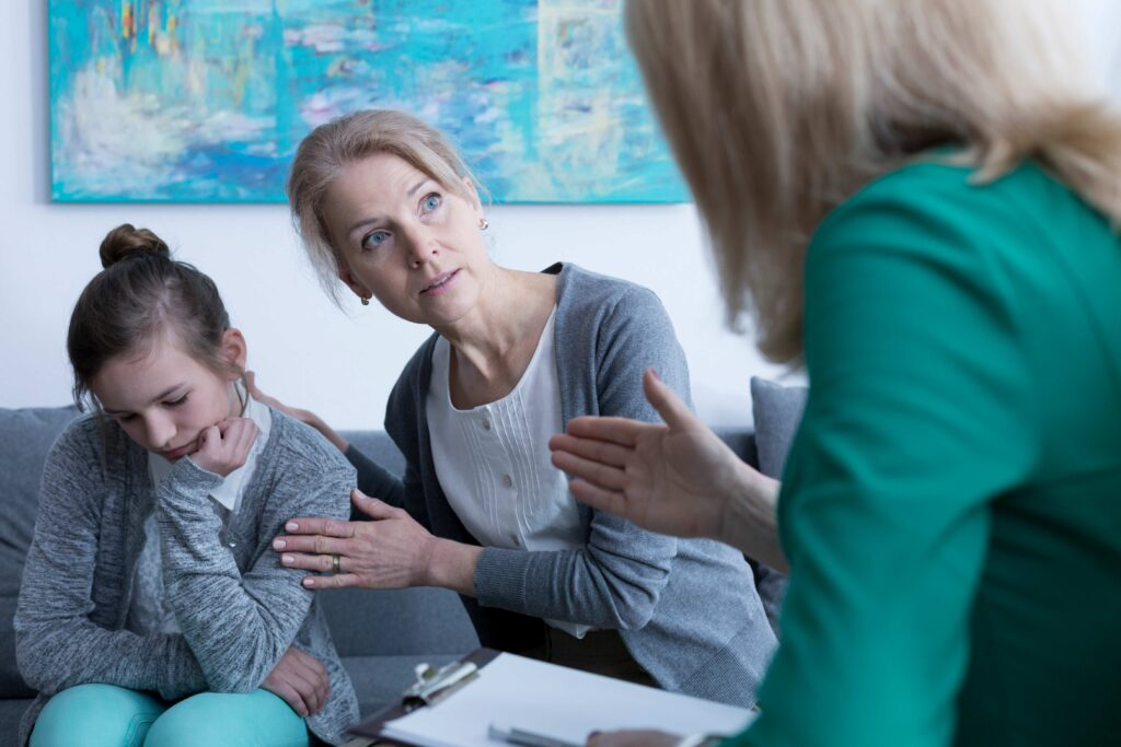 Concerned mom of special needs attentively speaks with a therapist during a session with her autistic daughter who appears withdrawn — capturing the emotional weight of advocacy, behavioral therapy, and navigating mental health challenges in special needs parenting.