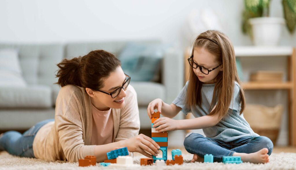 Joyful mom of special needs builds colorful blocks with her young autistic daughter on the floor — illustrating therapeutic play, developmental milestones, and the magic of one-on-one time that fosters confidence and cognitive growth in neurodiverse children.