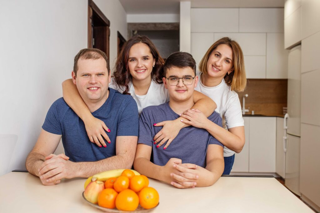 Supportive mom of special needs stands proudly behind her teenage autistic son, along with the whole family gathered in their kitchen — a powerful representation of unconditional love, autism acceptance, and daily life in a neurodiverse household.