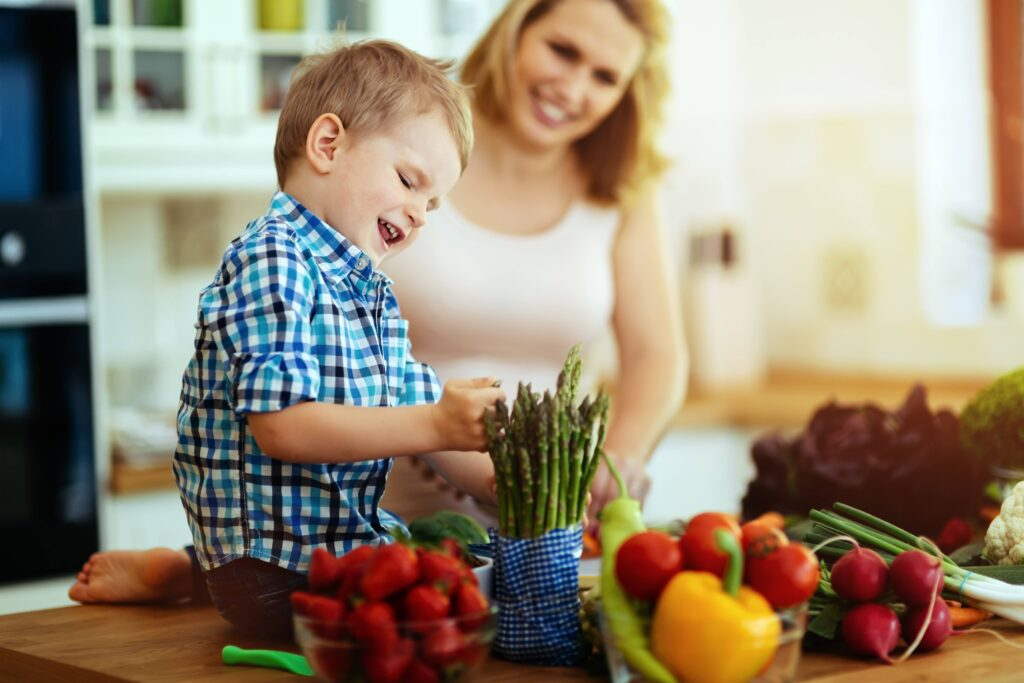 Young autistic boy happily helps his mom of special needs in the kitchen, learning to identify colorful vegetables during sensory-based cooking play — supporting motor skills, independence, and everyday learning through hands-on activities for neurodivergent children.