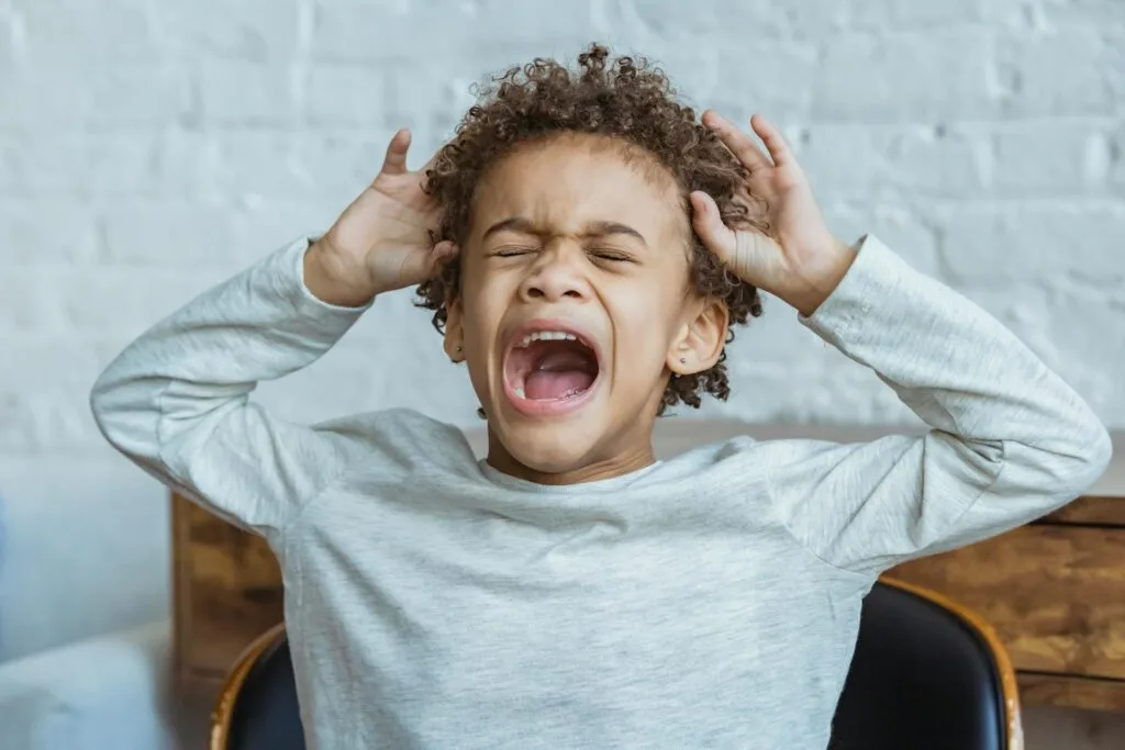 Child in the middle of a meltdown, hands covering ears and mouth open, showing the overwhelming sensory experience that distinguishes meltdowns from tantrums in special needs children